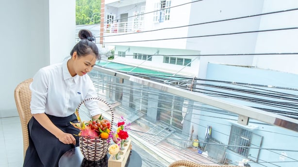 A woman sitting on a balcony with wicker chairs and a table, a basket of flowers, and a view of a building and greenery. - TiMee Hotel & Apartment (Nam Nha Trang)