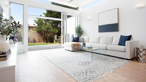 A modern living room with a white sofa, a glass coffee table, and a large window overlooking a backyard.