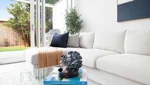 A modern living room with a white sofa, a glass coffee table with a book and a plant, and a view of a backyard with a wooden fence and greenery.