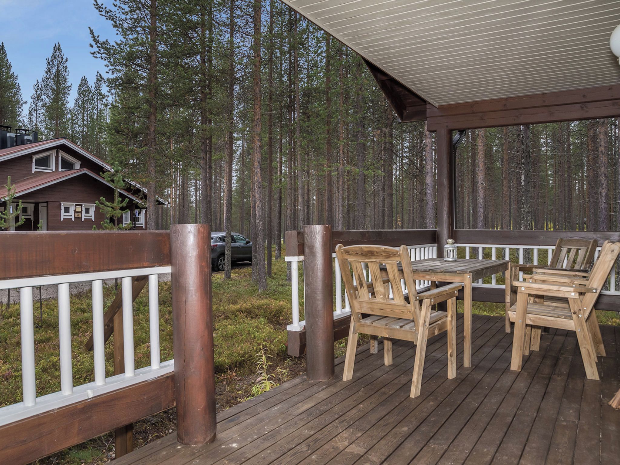 Eine Holzterrasse mit Tisch und Stühlen, umgeben von einem weißen Geländer, mit Blick auf einen Wald.