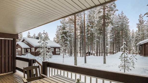 A wooden deck with a table and chairs, overlooking a snowy forest with cabins. - Albatrossi by Interhome (Kittilä)