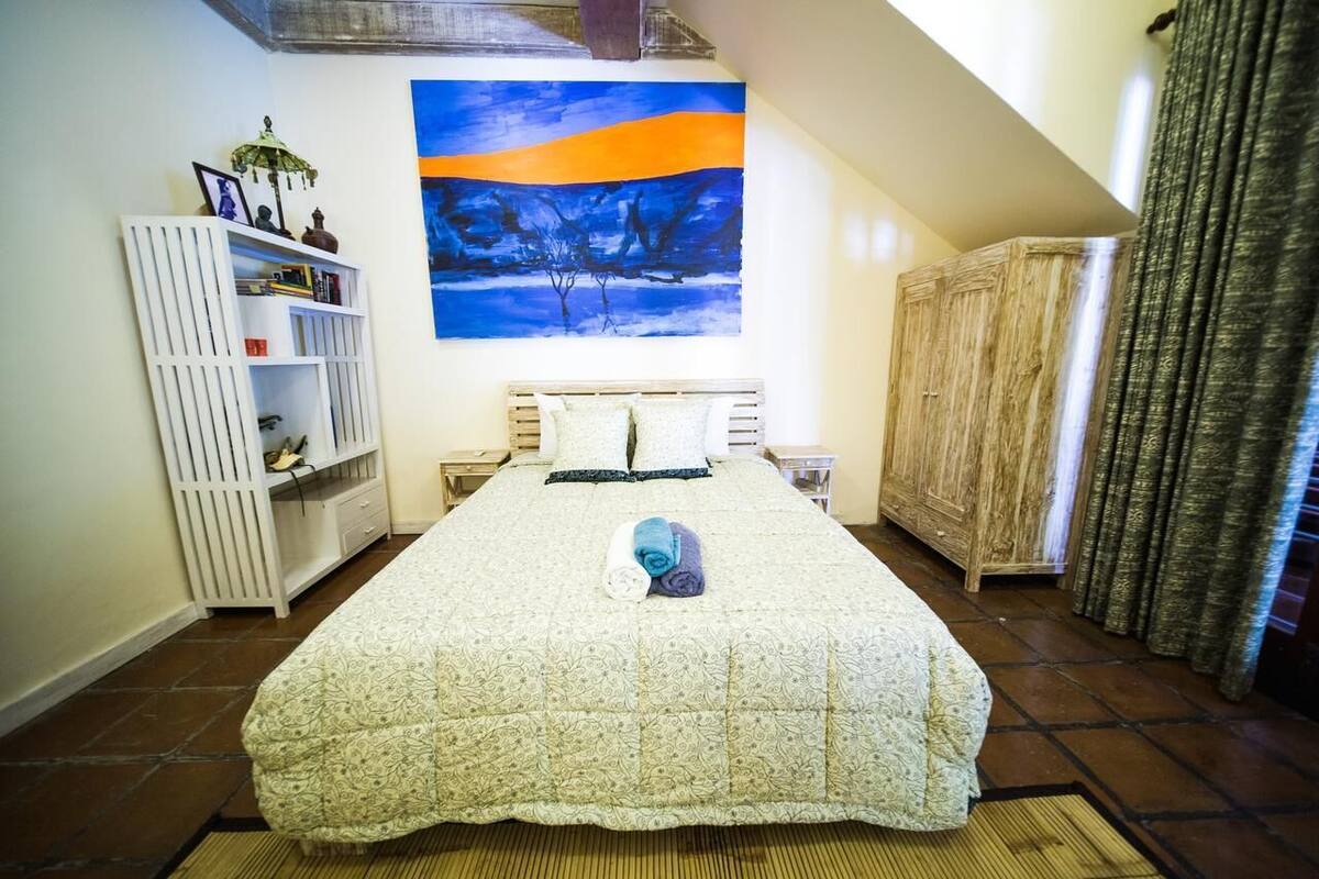 A bedroom with a large bed, a white wardrobe, a bookshelf, and a painting on the wall.
