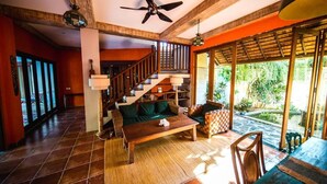 A spacious living area with a wooden staircase, a sofa, a coffee table, and a view of a garden through large windows. - Easy Surf Villa (Pererenan)