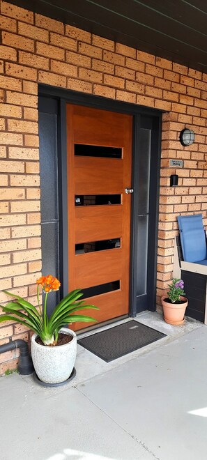 A brick building with a wooden door, a potted plant with orange flowers, and a small sign on the wall. - Quality studio style accommodation in the heart of the beautiful Tamar Valley (Beaconsfield)