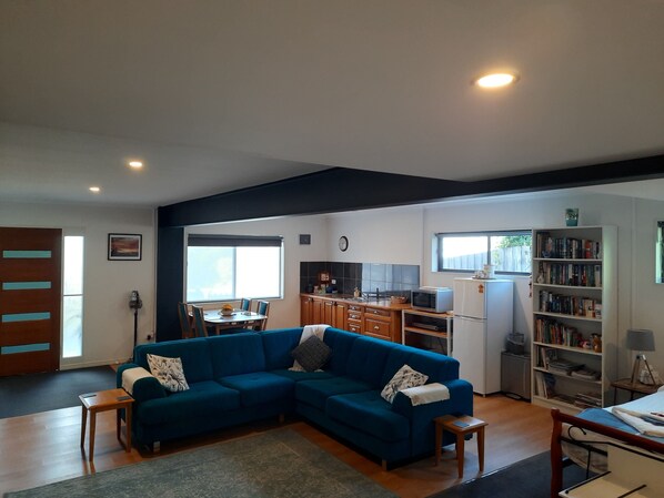 A living room with a blue sectional sofa, a dining table with chairs, a kitchen area with cabinets and appliances, and a bookshelf.
