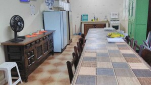 Un coin repas avec une longue table, des chaises, un ventilateur, un réfrigérateur et un meuble vert.