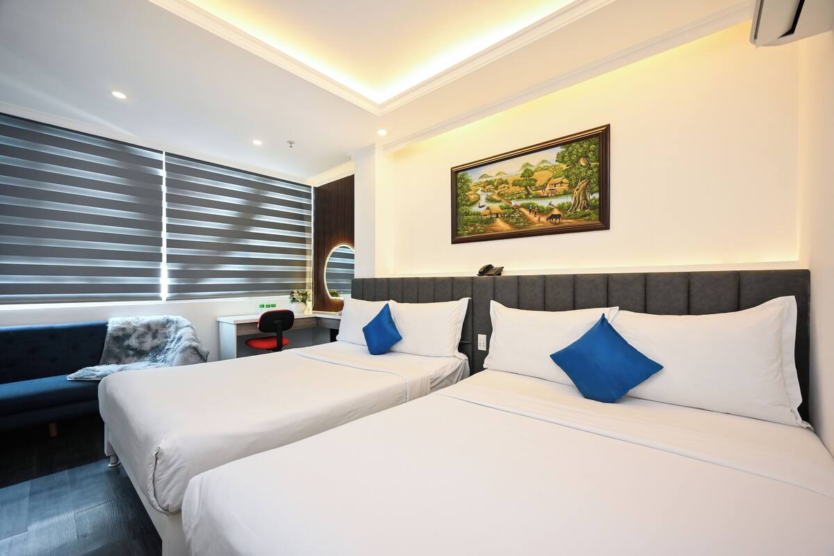 Une chambre d’hôtel avec deux lits, un bureau, une chaise et une grande fenêtre avec des stores.