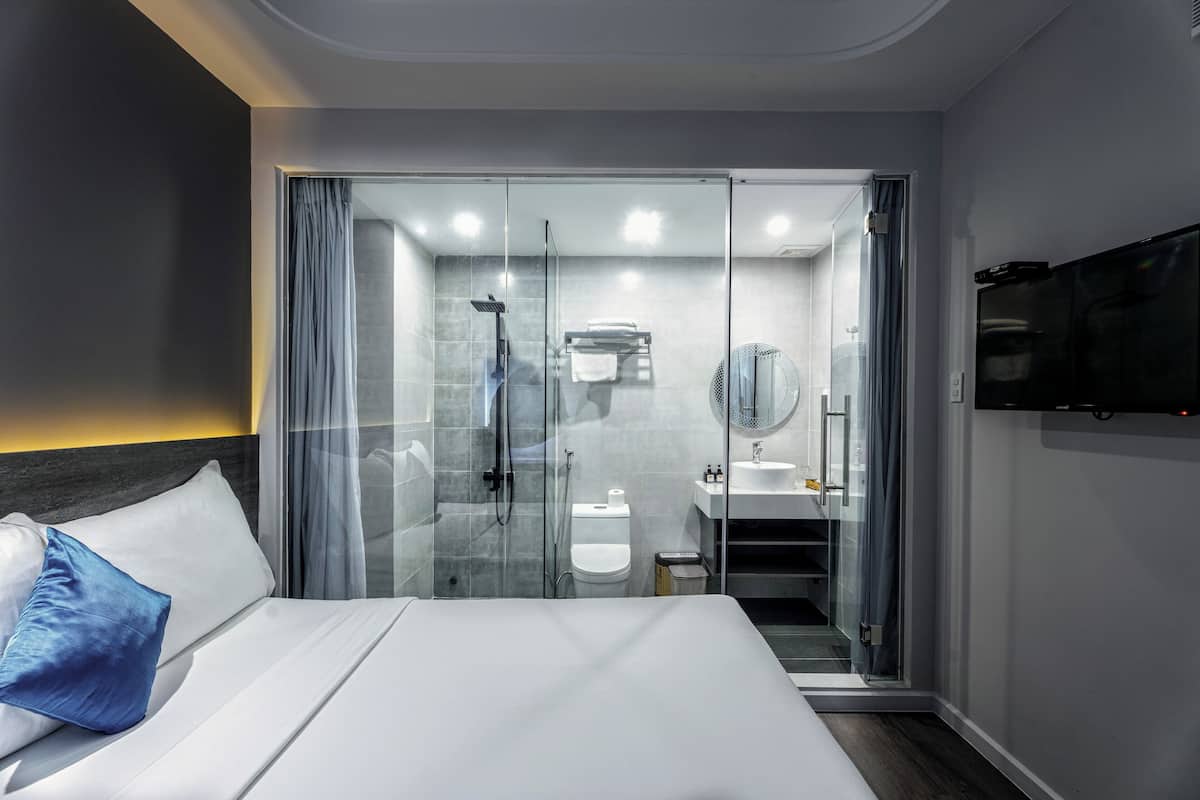 Une chambre d’hôtel moderne dotée d’un grand lit, d’une douche séparée par des parois en verre, d’un téléviseur fixé au mur et d’une salle de bain ouverte avec un lavabo et un miroir.
