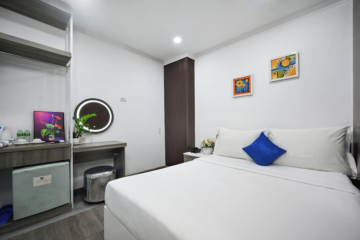 Une chambre d’hôtel moderne équipée d’un lit, d’un bureau, d’un petit réfrigérateur, d’une poubelle, d’un miroir et de deux œuvres d’art accrochées au mur.