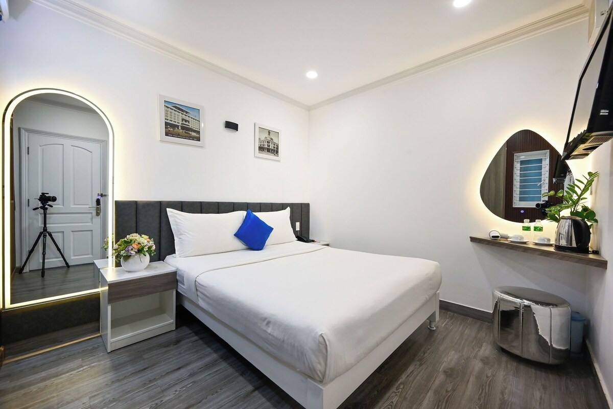 Ein modernes Hotelzimmer mit Bett, Nachttisch, Spiegel und einem kleinen Schreibtisch mit Kamera auf Stativ.