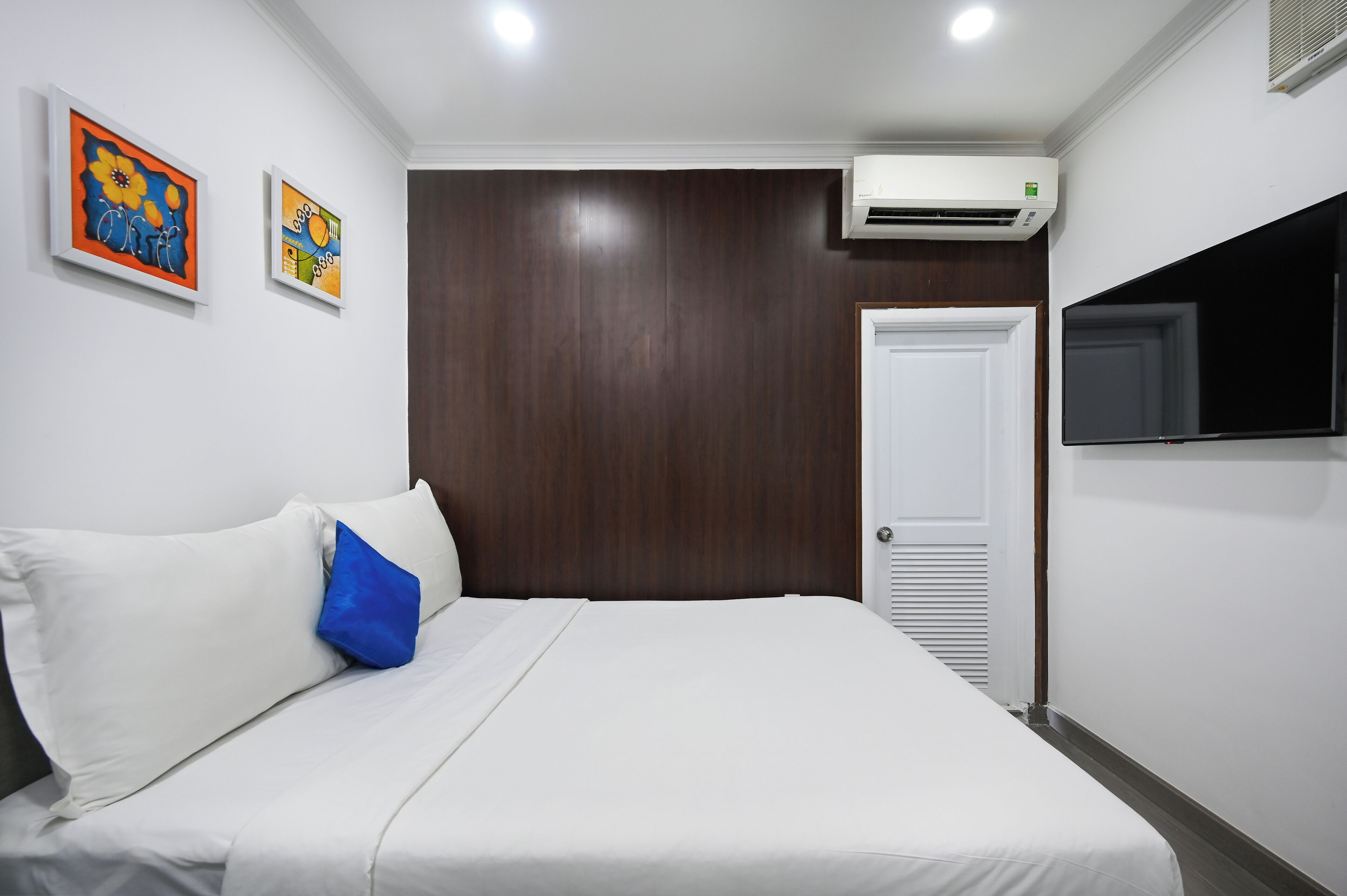 A hotel room with a bed, white bedding, a blue pillow, a wall-mounted TV, and two paintings on the wall.