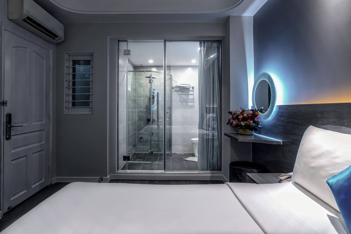 Une chambre d’hôtel moderne comprenant un lit, une douche en verre, une coiffeuse avec un miroir et un petit coin salon.