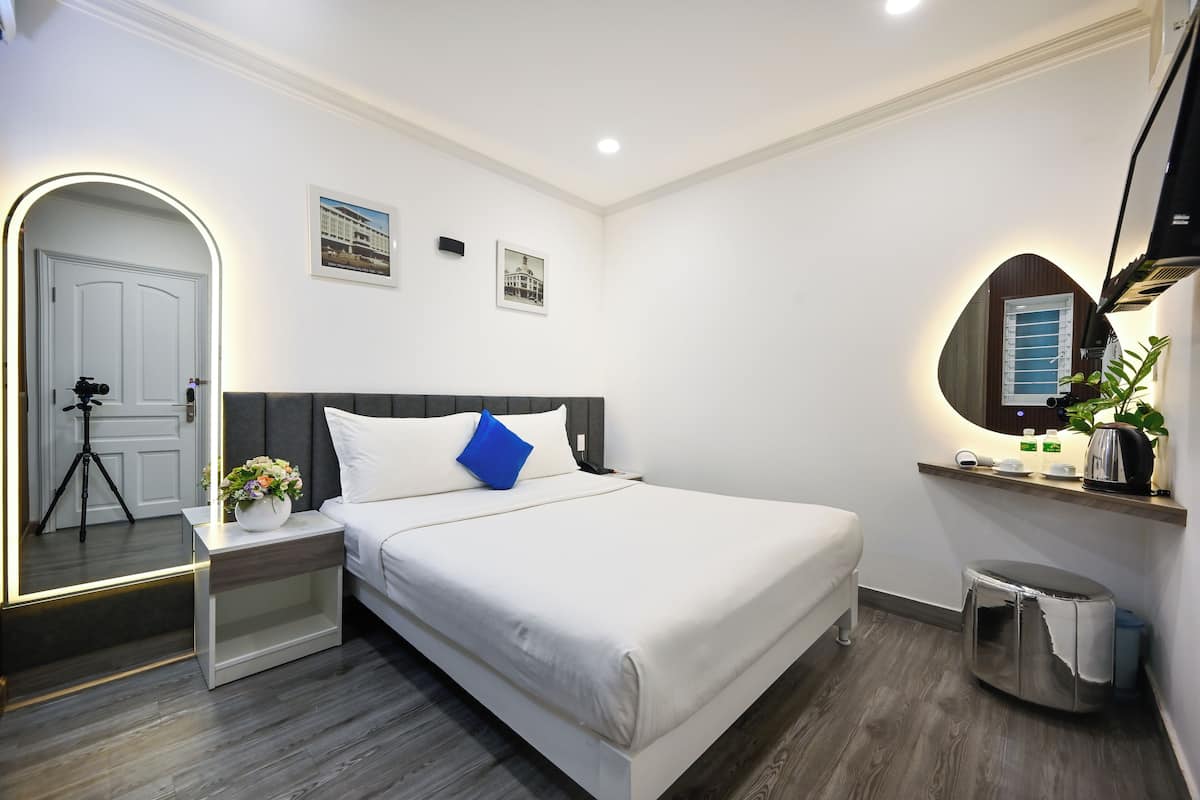 Une chambre d’hôtel moderne avec un grand lit, une table de chevet ornée d’un vase de fleurs, un miroir, un petit tabouret et une étagère contenant des articles de toilette.