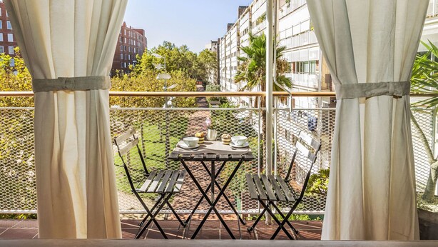 Un balcón con mesa y sillas, con vistas a un jardín y edificios.