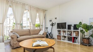 Una sala de estar con un sofá color crema, una mesa de centro redonda de madera con un libro abierto y vasos, un televisor, parlantes y una estantería con libros y una planta.