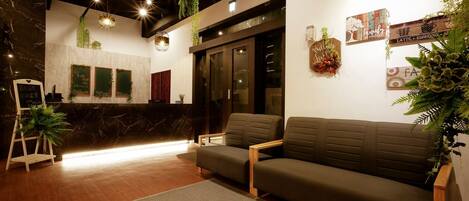 Una zona de recepción moderna con mostrador de mármol, sillas de madera y un sofá. Se observan decoraciones en las paredes y plantas.
