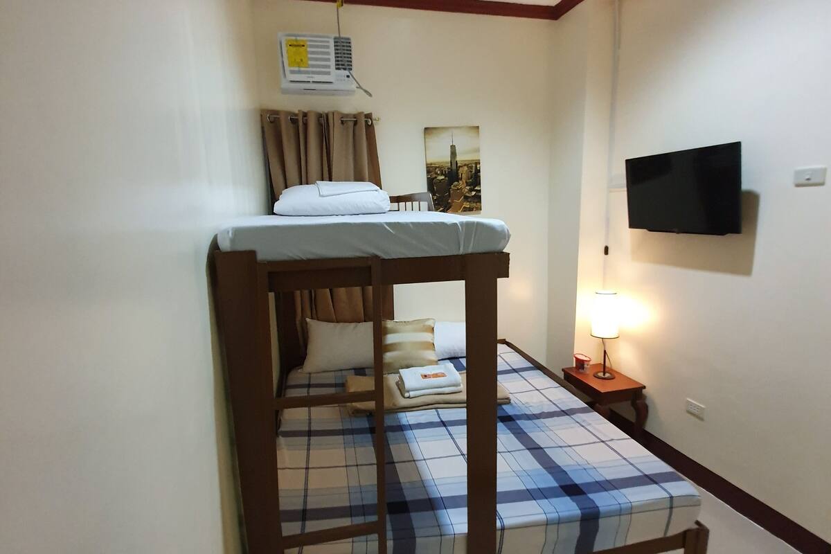 A single bed with a checkered bedspread, a bedside lamp, a flat-screen TV mounted on the wall, and a window with curtains.