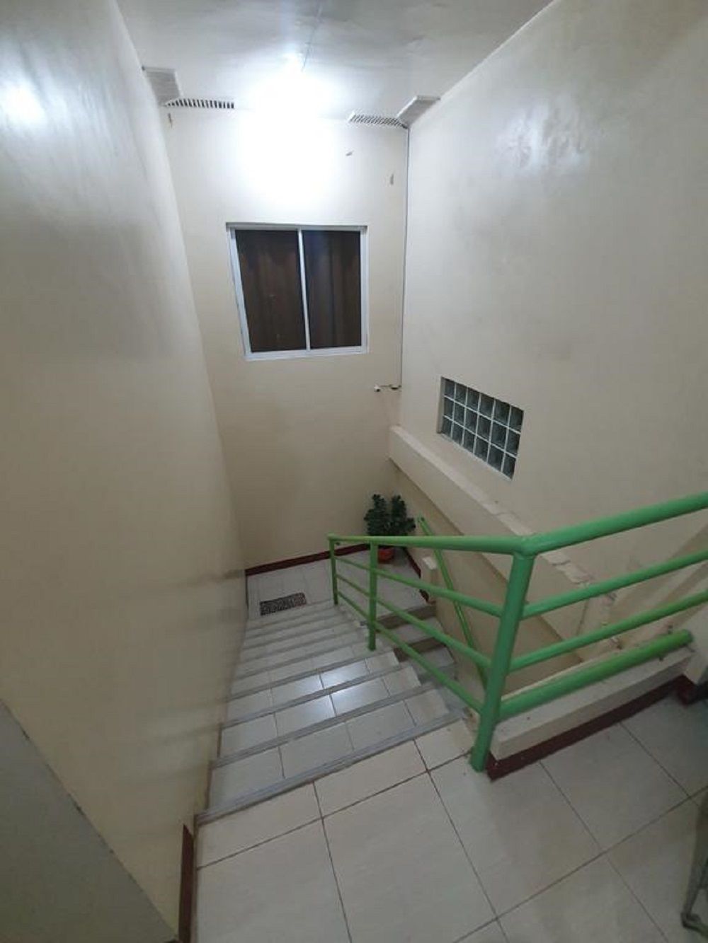 Una escalera estrecha con barandilla verde, paredes blancas y un pequeño ventanal en la parte superior.