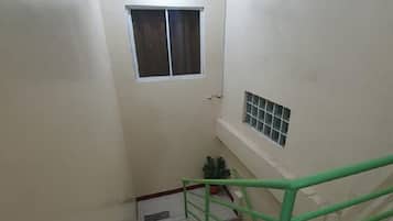 Una escalera estrecha con barandilla verde, paredes blancas y un pequeño ventanal en la parte superior.