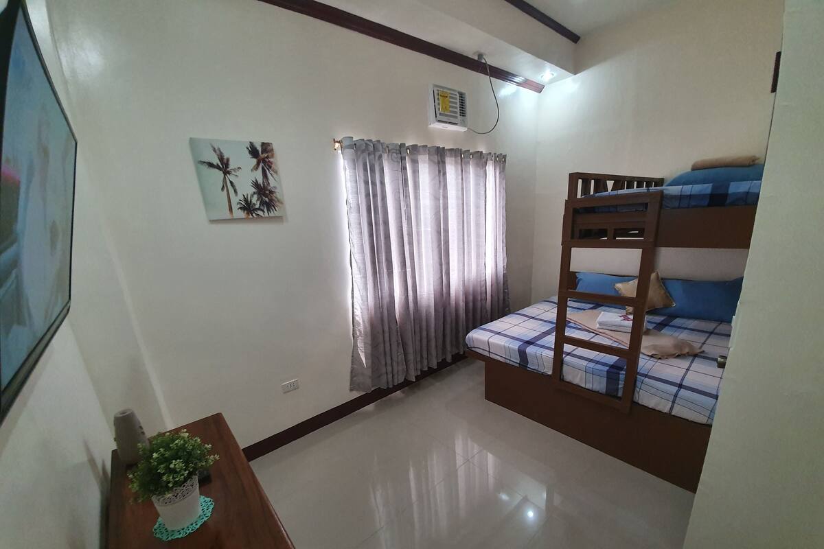 A small, well-lit room with a bunk bed, a desk with a plant, and a painting on the wall.