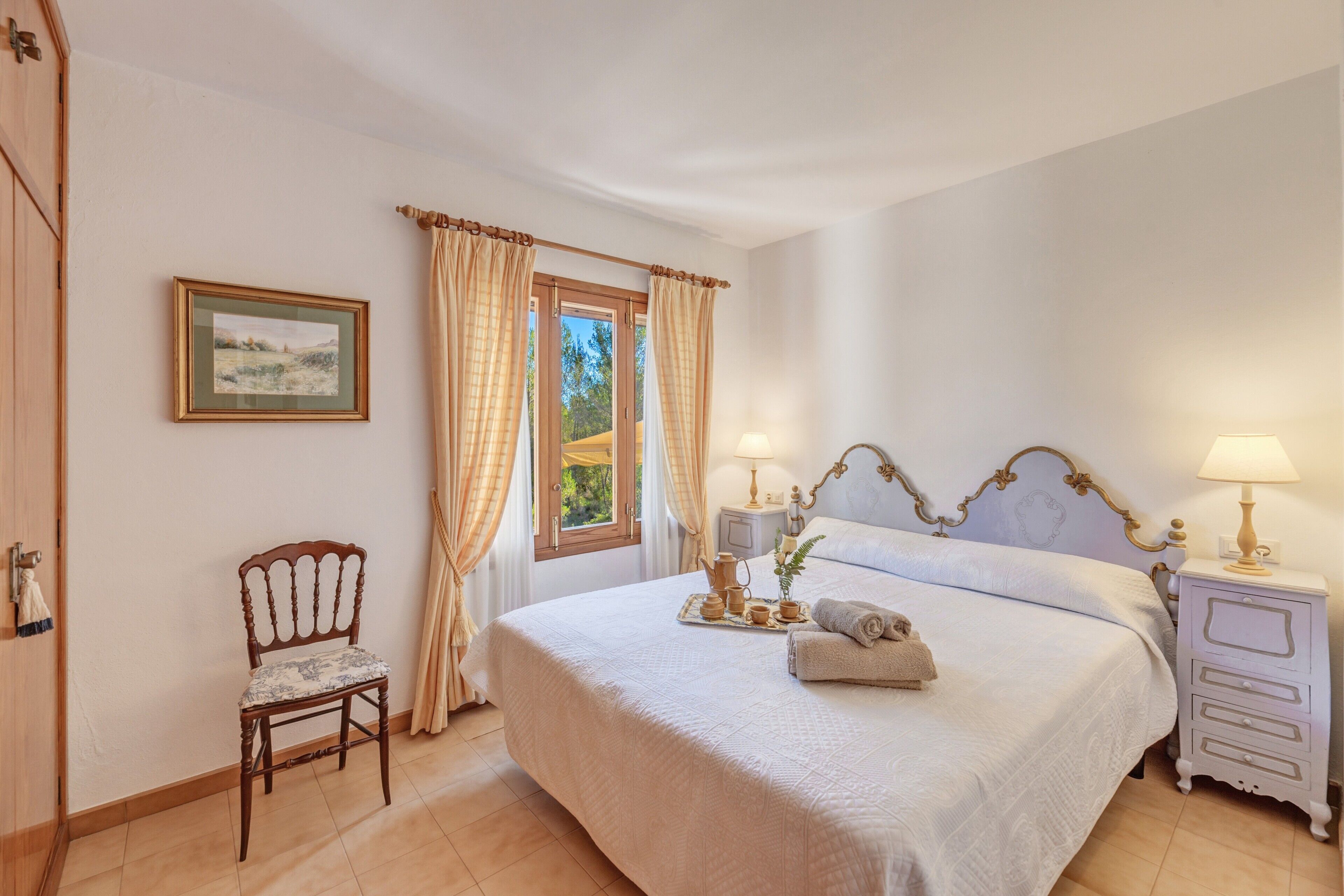 A bedroom with a bed, a chair, a bedside table with lamps, a window with curtains, and a framed picture on the wall.