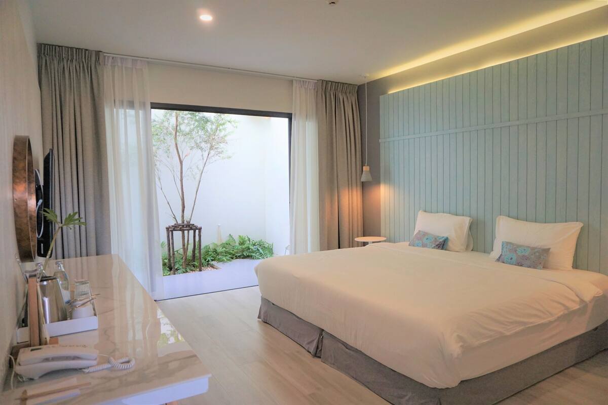 A modern hotel room with a large bed, a bedside table with a phone and a lamp, and a view of greenery outside.