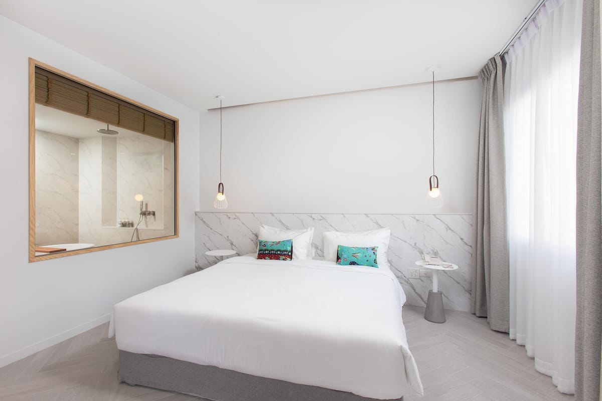 Un dormitorio moderno con una cama grande, una mesita de noche, un espejo y un baño visible a través de una puerta corredera.