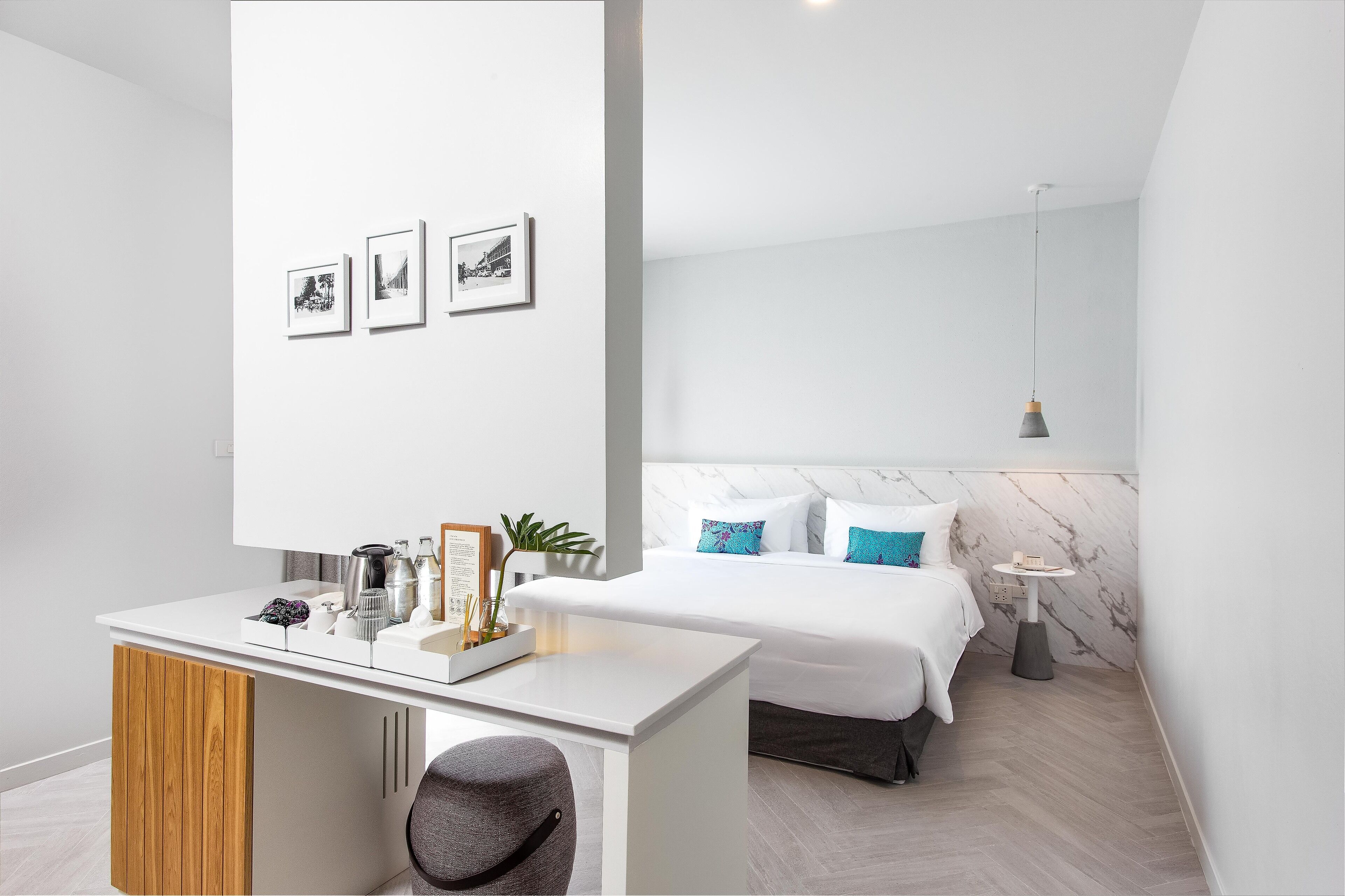 Ett modernt hotellrum med en vit säng, ett litet skrivbord, en stol, en sänggavel i marmor och en vägghylla med inramade bilder.
