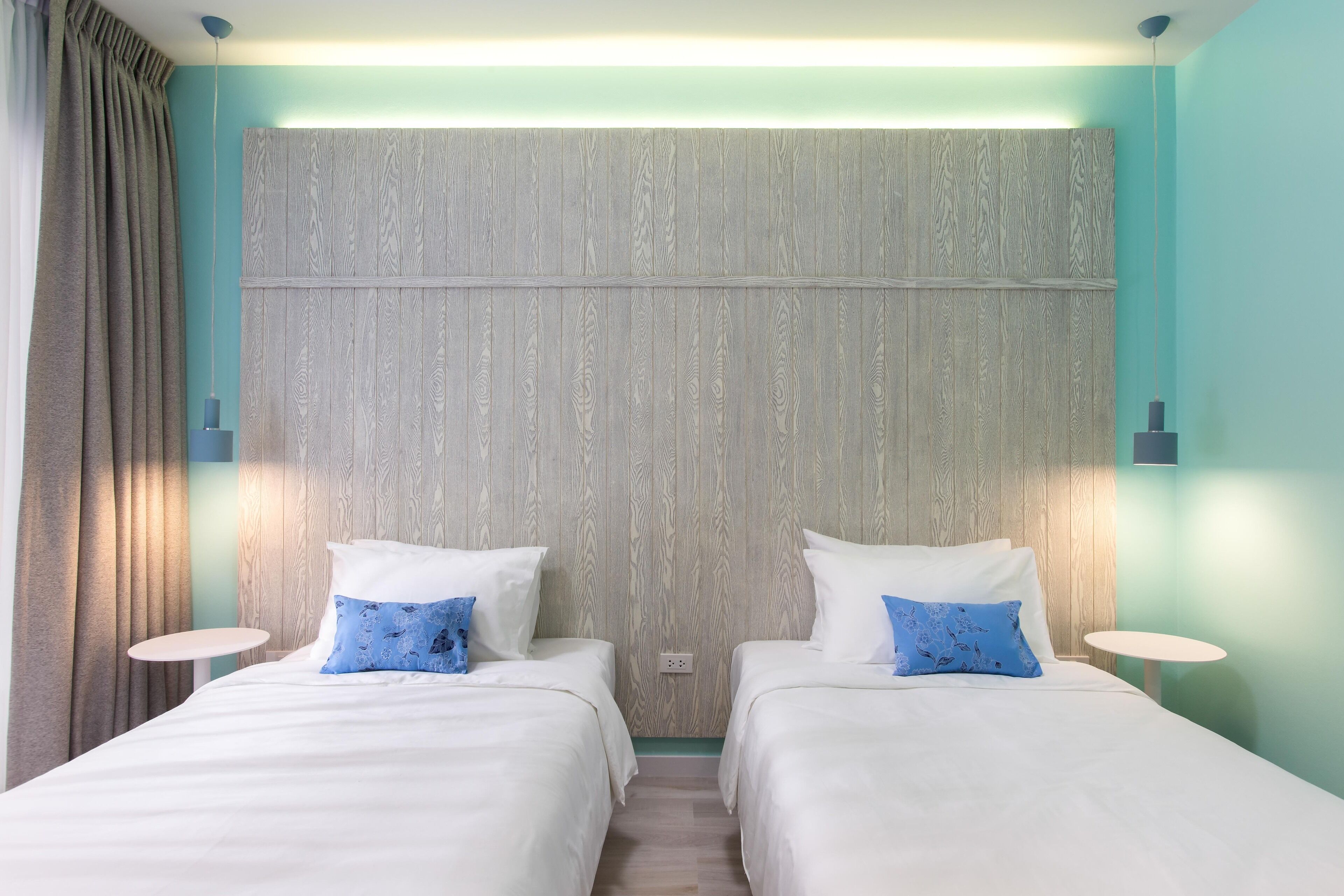 Habitación de hotel con dos camas, cabecero de madera, paredes color turquesa y una pequeña mesa redonda.