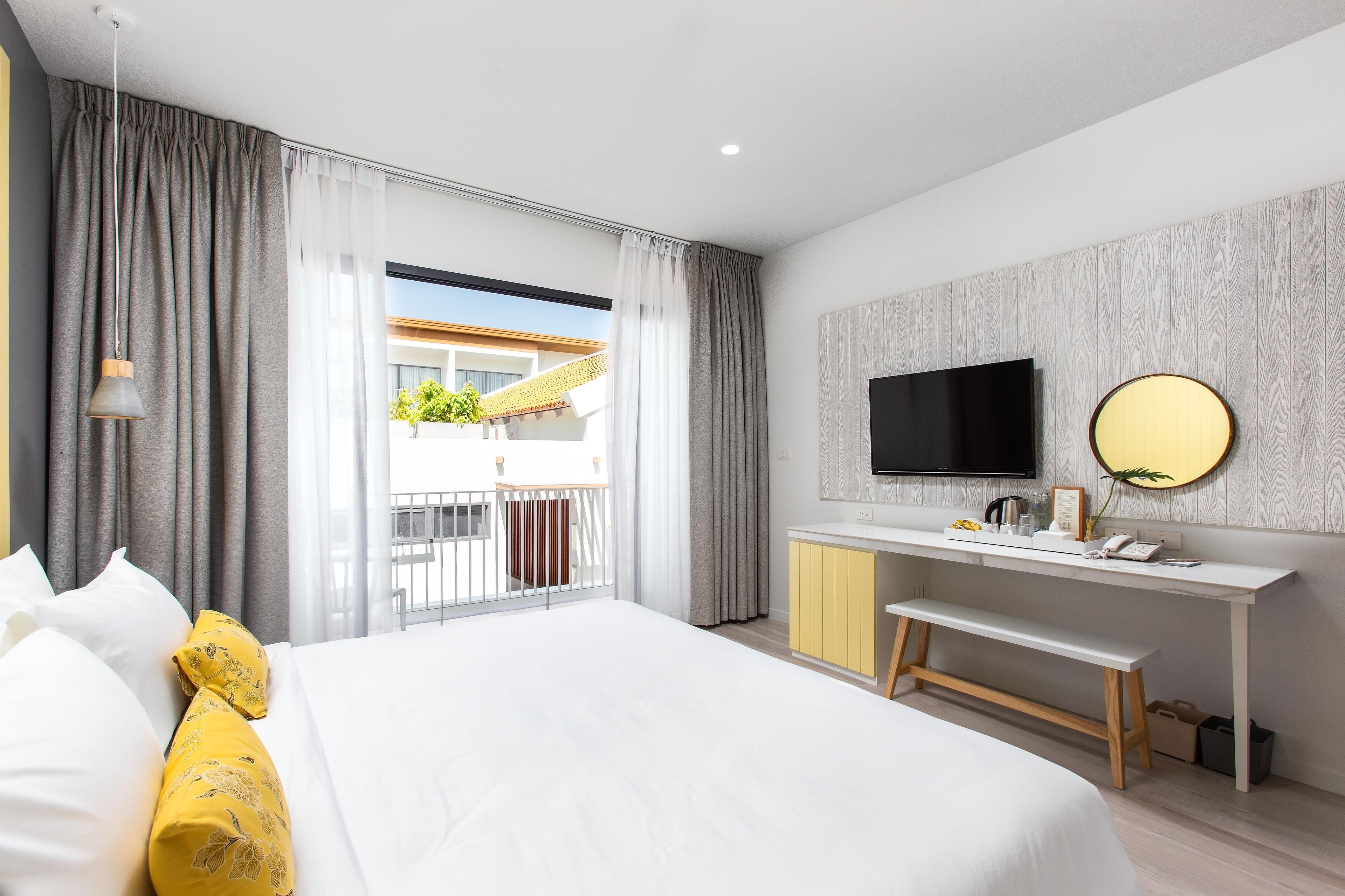 A modern hotel room with a large bed, a desk with a flat-screen TV, a bench, and a balcony with a view of the outside.