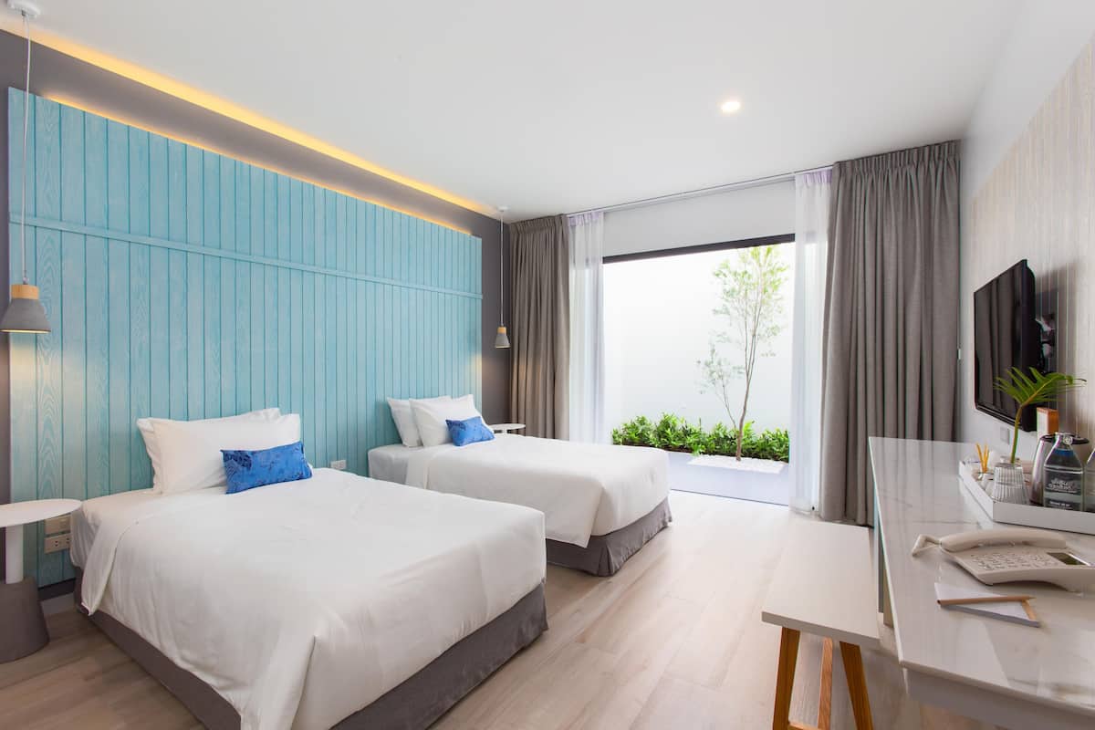 A modern hotel room with two beds, a desk, a TV, and a large window with a view of greenery.