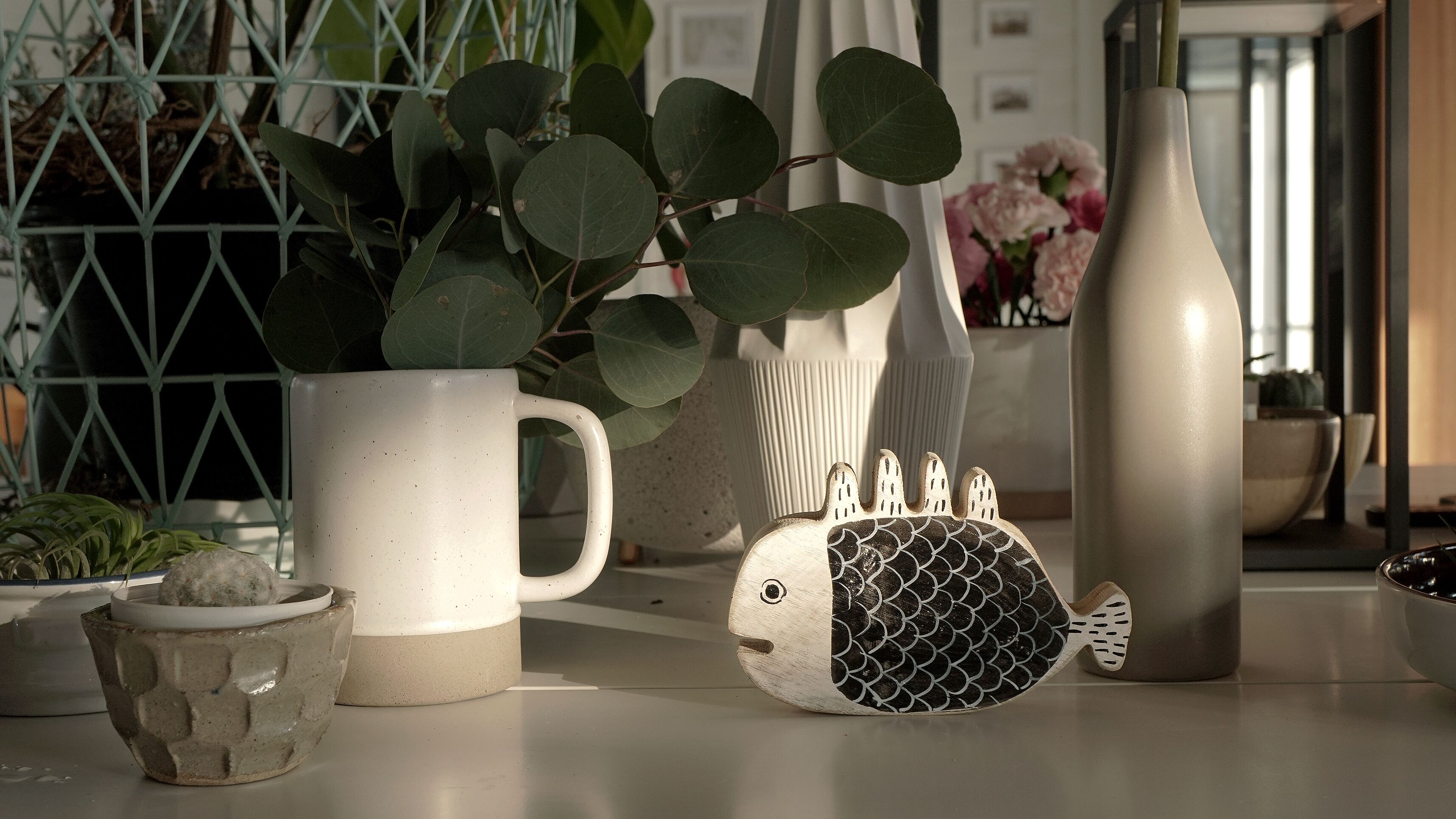 A kitchen counter with a white mug, a ceramic fish, a small plant, and a vase with flowers.