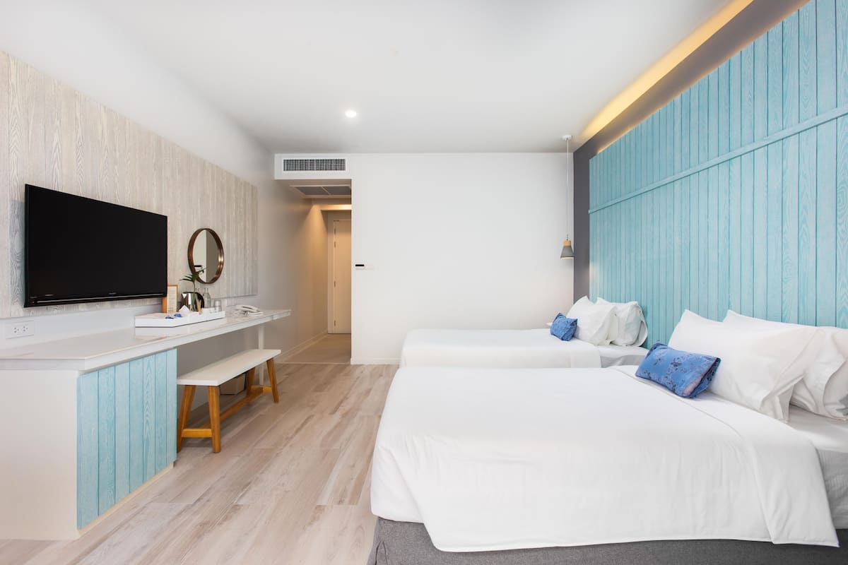 Habitación de hotel con televisor de pantalla plana, escritorio con espejo, dos camas con ropa blanca y una pared de acento azul.