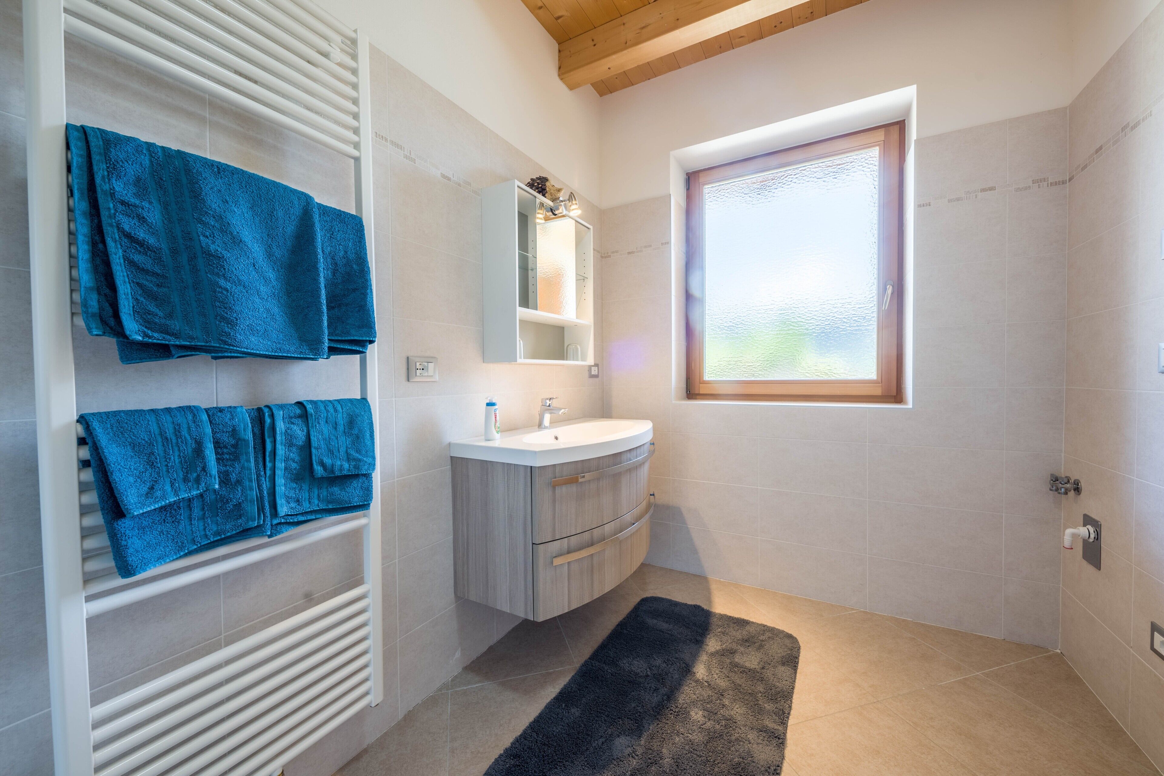 Un baño con un tocador blanco, un perchero con toallas azules, un tapete negro y una ventana con vistas.