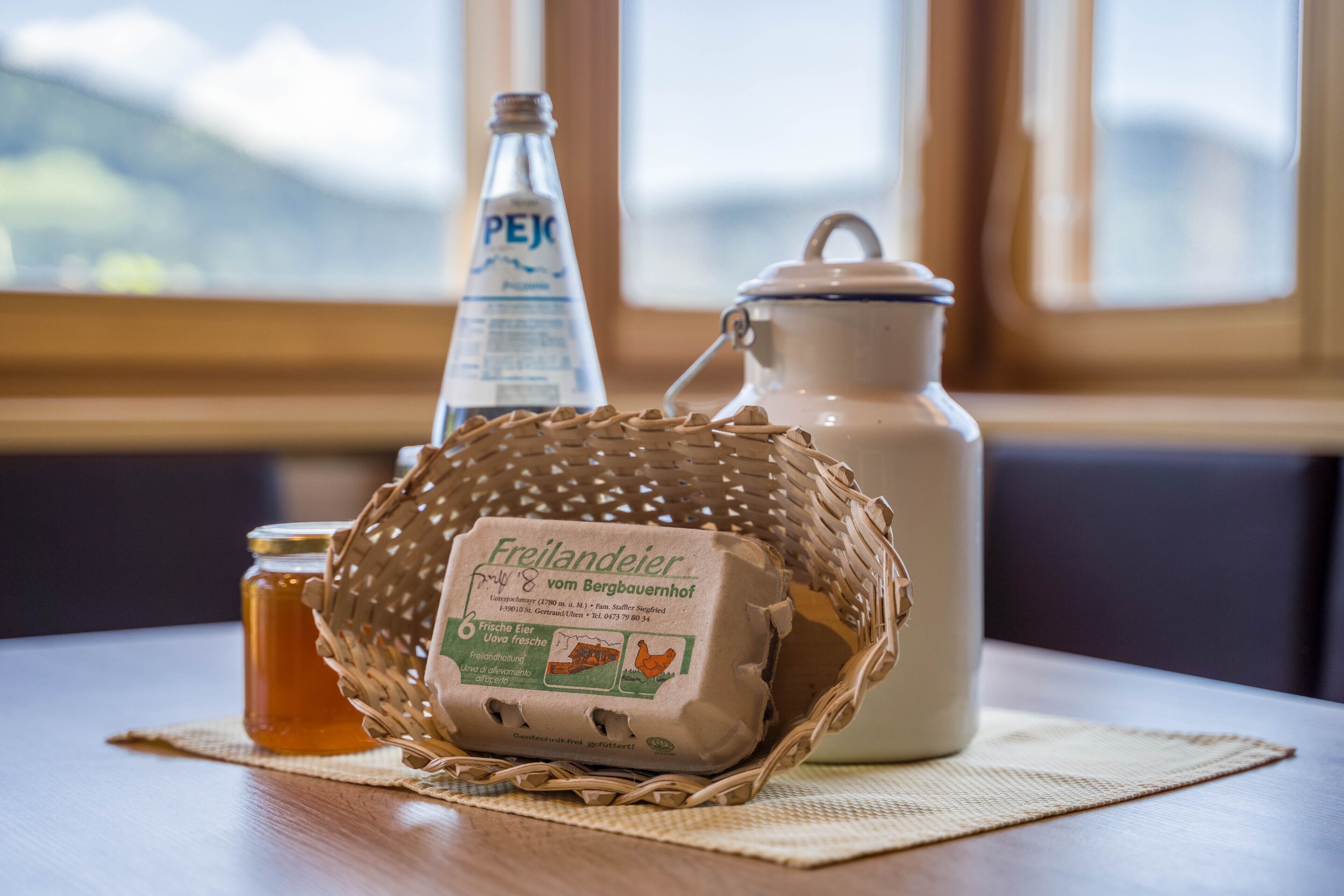 A table with a woven basket containing a package of eggs, a bottle of PEJ beer, and a ceramic pitcher.