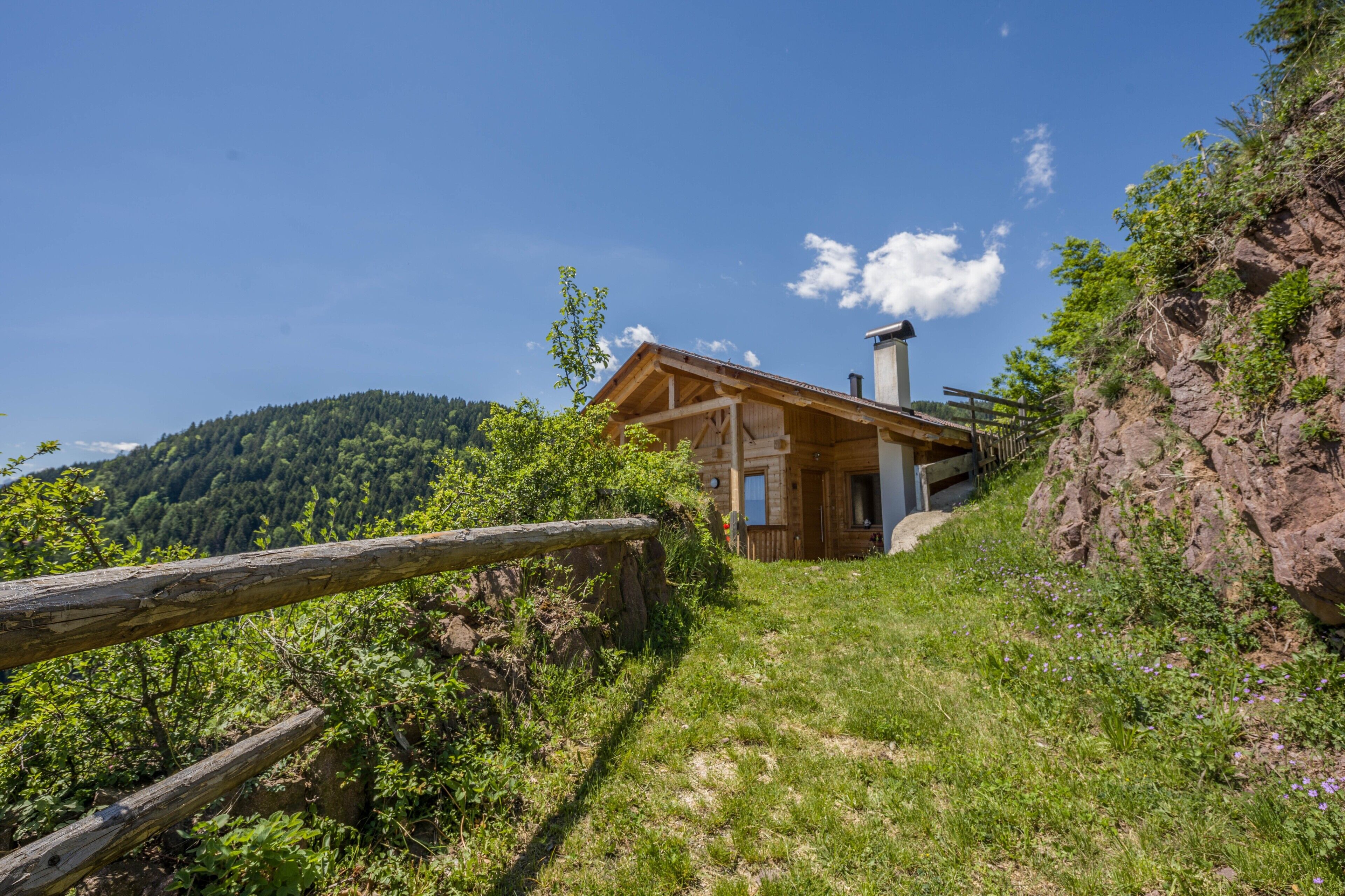 A wooden cabin with a chimney, surrounded by a forested hillside.