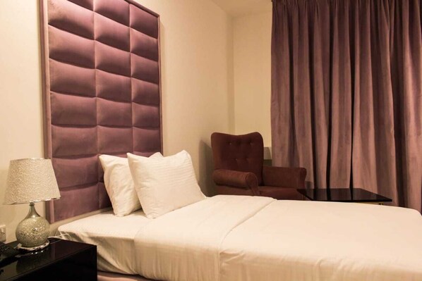 A hotel room with a tufted headboard, a bed with white bedding, a brown armchair, a small table, and a lamp on a nightstand.