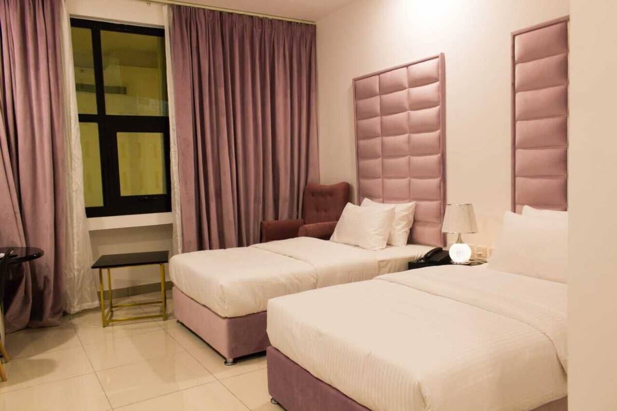 A hotel room with two beds, a window with curtains, a bedside table with a lamp, and a chair.