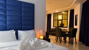 A hotel room with a large bed, a dining area with a table and chairs, and a view of the city at night. - Parkside Plaza Hotel - Muscat (Muscat)
