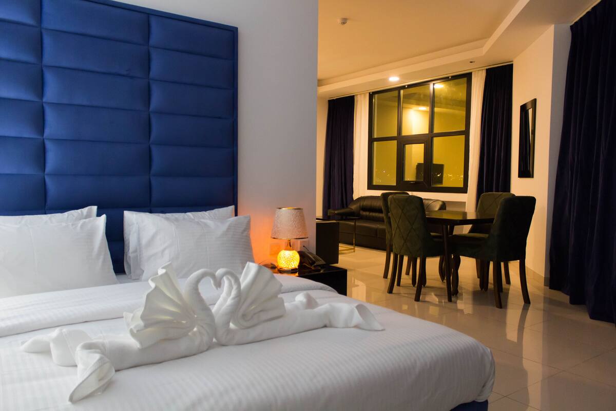 A hotel room with a large bed, a dining area with a table and chairs, and a view of the city at night.