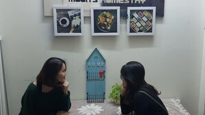 Una habitación con un cartel en la pared que dice 'Moon's Homestay' y tres cuadros con imágenes de comida. Se ve una puerta azul con un letrero que dice 'HOGAR'.