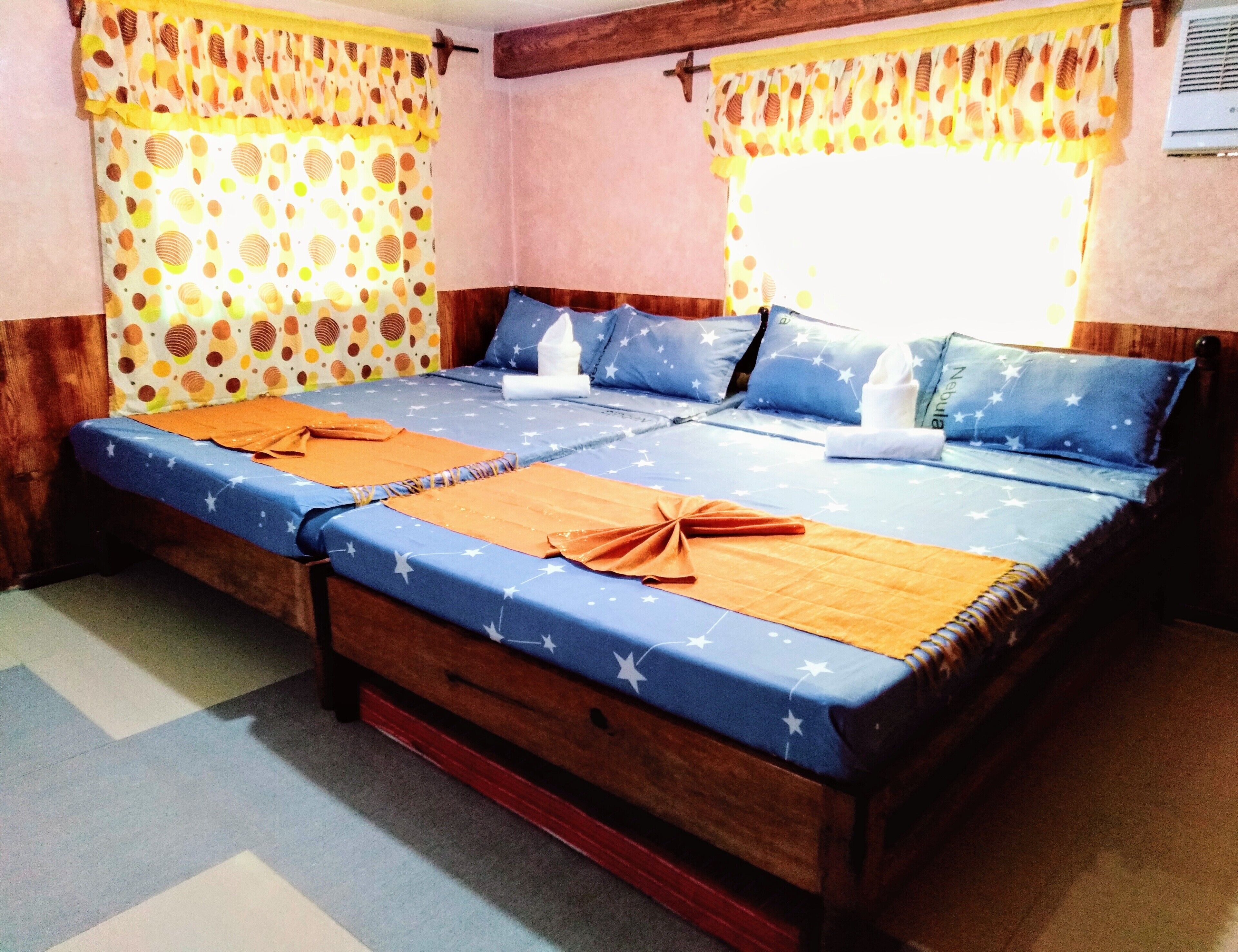 Una cama doble con una colcha azul y naranja estampada con estrellas, dos almohadas con diseños de estrellas y una cortina amarilla con lunares.