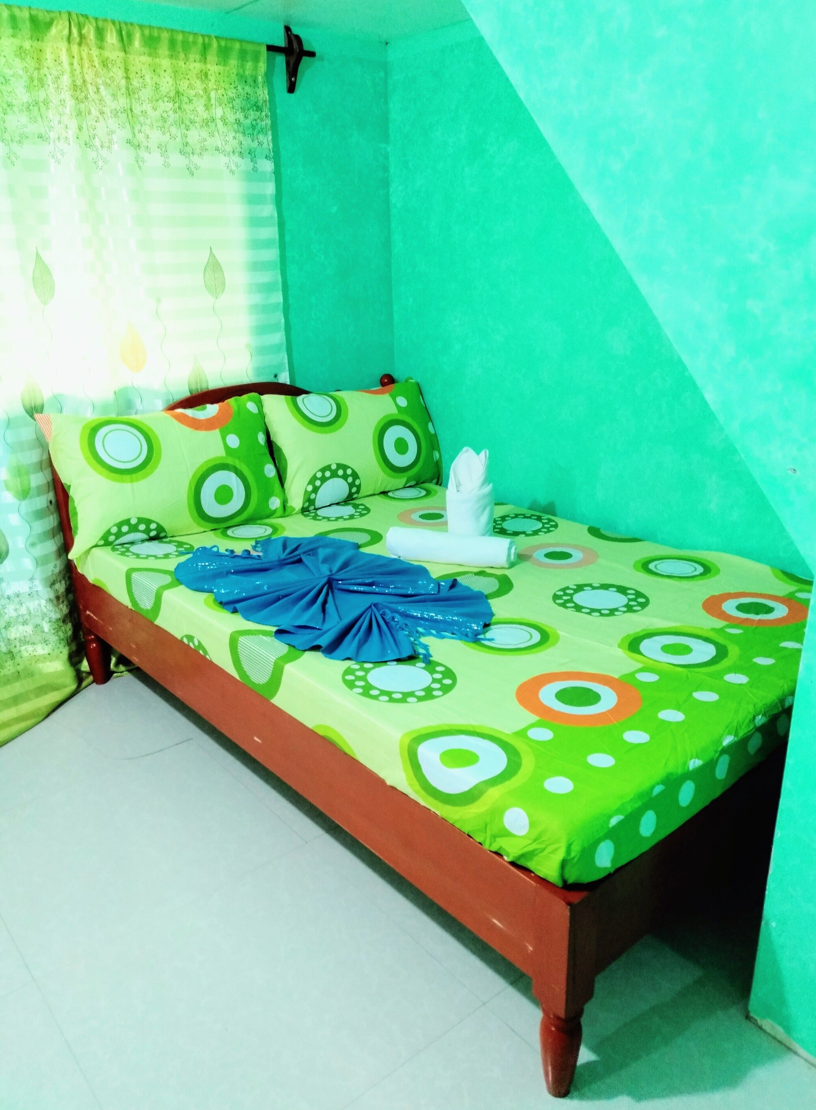 Una habitación pequeña con cama individual, colcha verde y blanca estampada, toalla azul y almohada con diseños circulares.