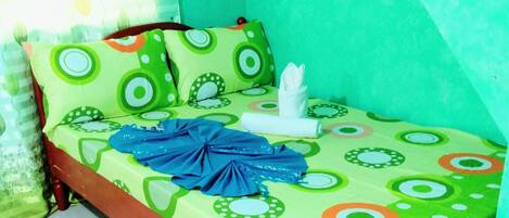 Una habitación pequeña con cama individual, colcha verde y blanca estampada, toalla azul y almohada con diseños circulares.
