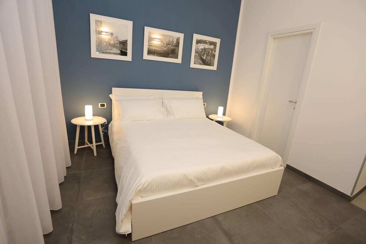 A neatly made bed with white bedding, two bedside lamps, and three black-and-white photographs on the wall.