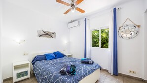 Un dormitorio con una cama, una mesita de noche, un ventilador de techo, un equipo de aire acondicionado, una ventana con cortinas y un adorno en la pared.
