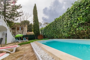 Piscina com borda branca, cercada por um arbusto, e um pequeno edifício com porta e janelas.