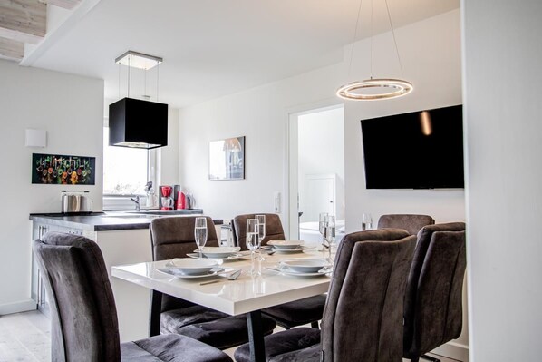 A modern dining area with a white table set for six, surrounded by dark upholstered chairs. A black pendant light hangs above the table, and a flat-screen TV is mounted on the wall to the right.