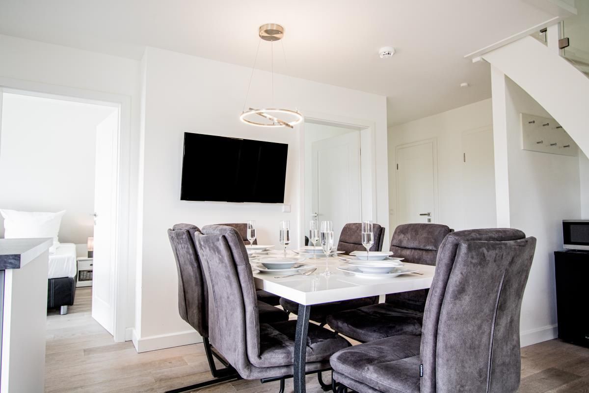 A modern dining area with a white table set for four, grey upholstered chairs, and a flat-screen TV mounted on the wall.