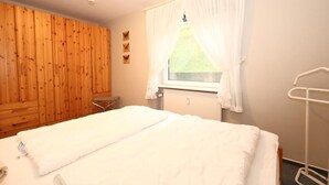 A bedroom with a wooden wardrobe, a bed with white bedding, a window with white curtains, and a white radiator. - Bangert House Cäcilia (Wenningstedt-Braderup (Sylt))